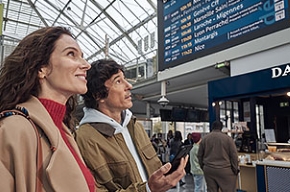 Photo of a man and a woman in a station in front of a screen displaying train departures and arrivals