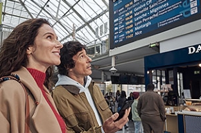 Foto van een man en een vrouw in een station voor een scherm met vertrekkende en aankomende treinen